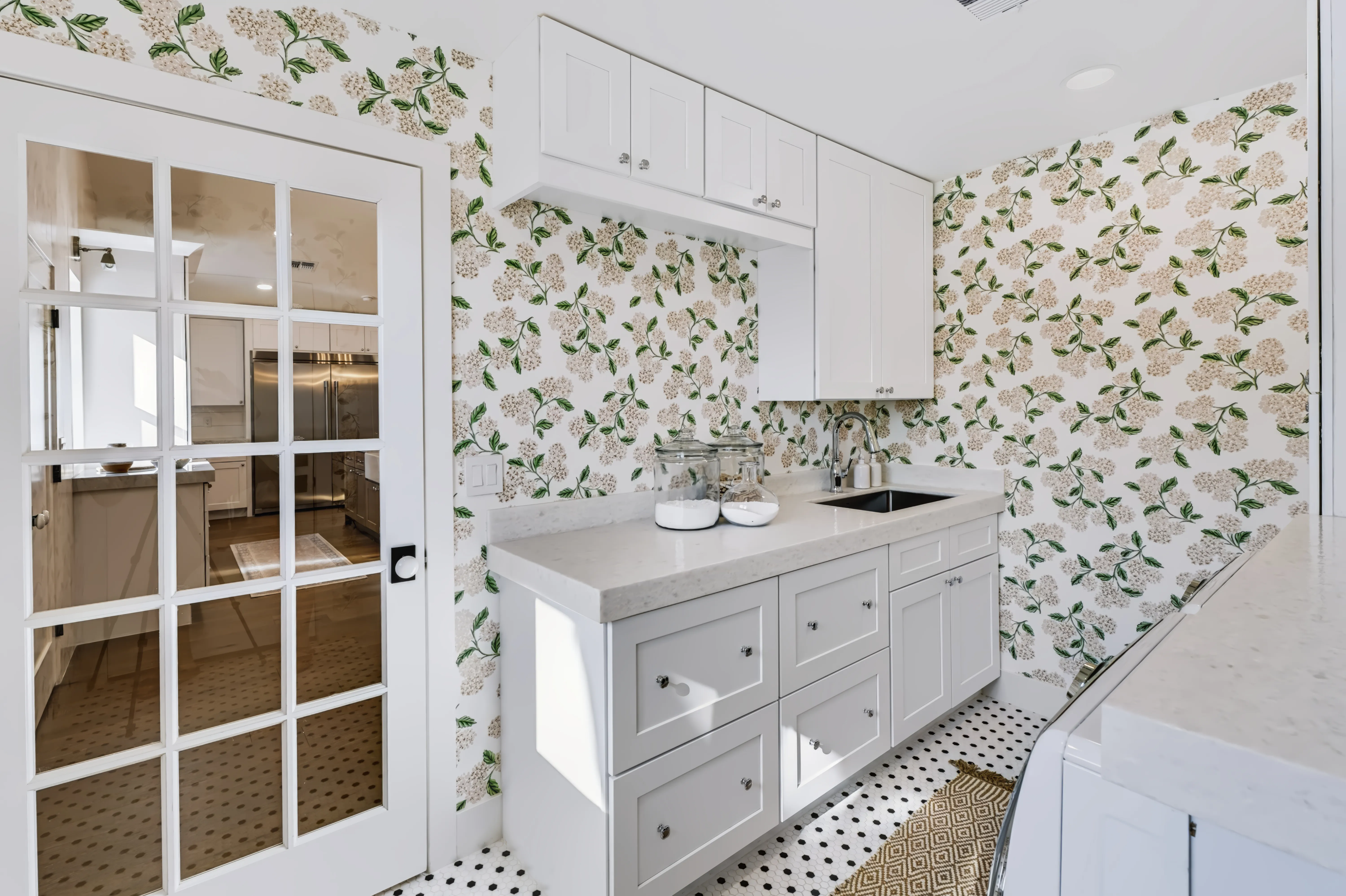 Laundry room papered in a hydrangea print, white cabinetry and a glass-paned door to the kitchen