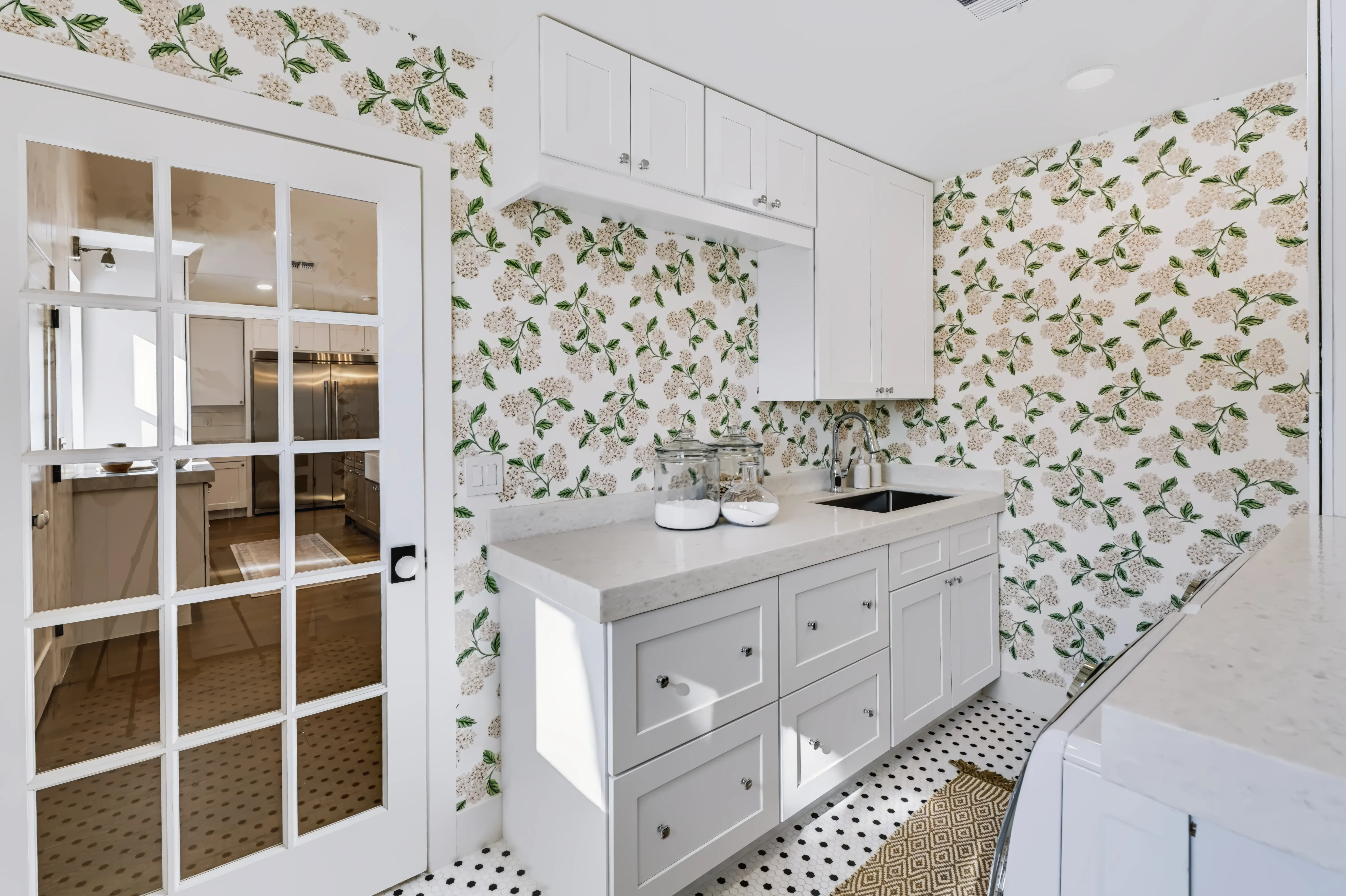 Laundry room papered in a hydrangea print, white cabinetry and a glass-paned door to the kitchen