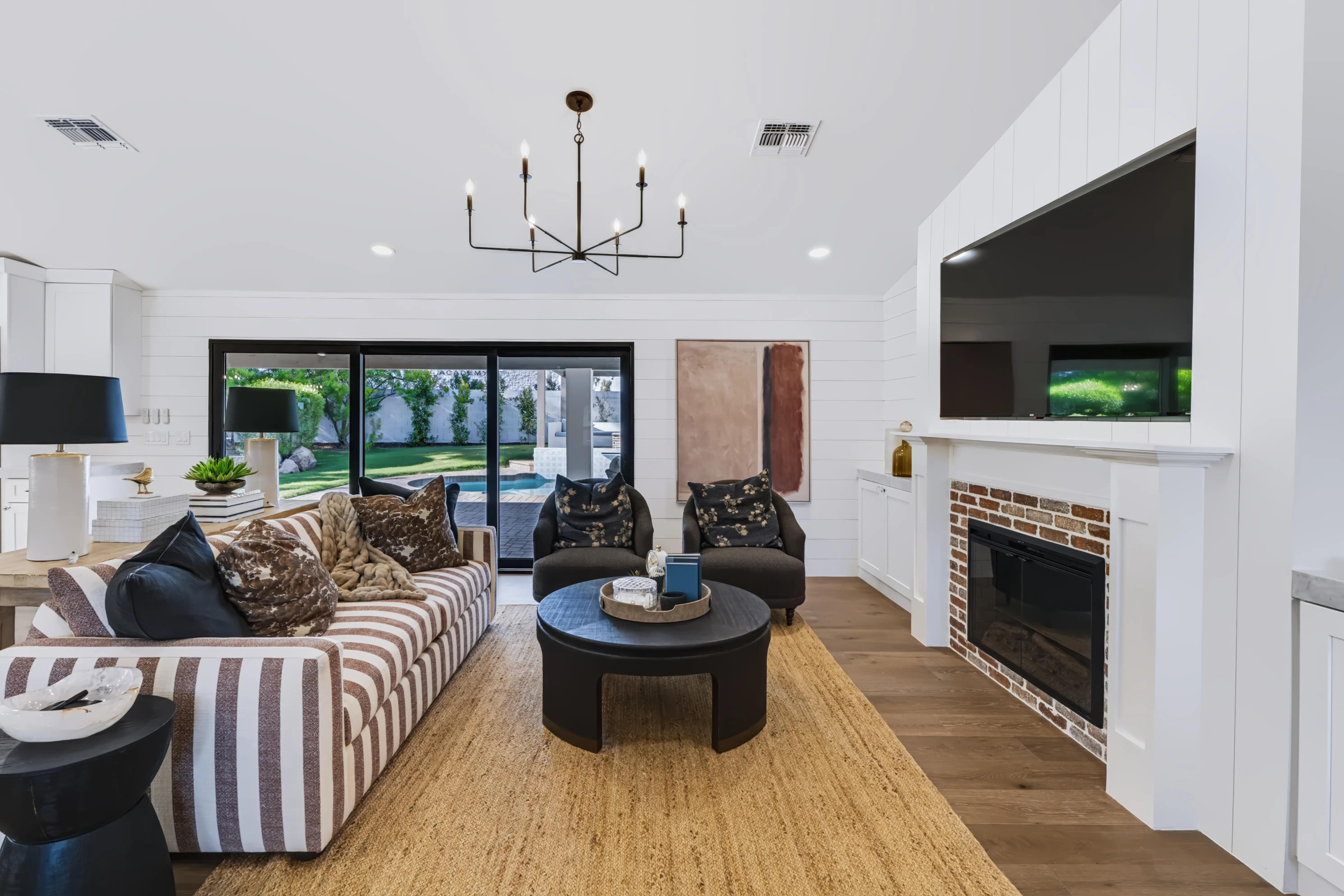 Great room with a whitewashed brick fireplace and mounted TV, a striped sofa and sliding glass to the pool