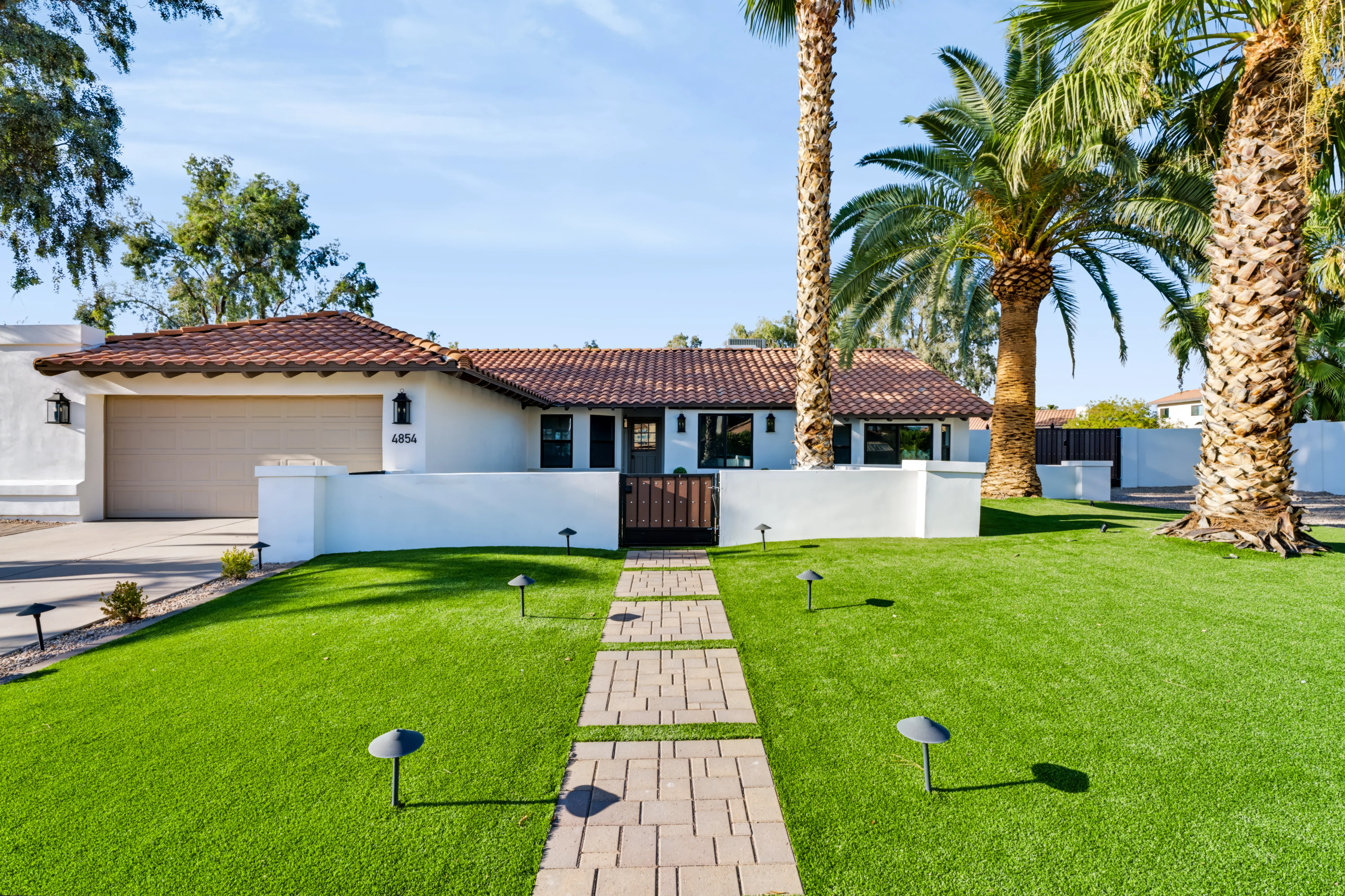 White Spanish ranch with a red barrel-tile roof, date palms and a paver walk across the lawn to a gated courtyard