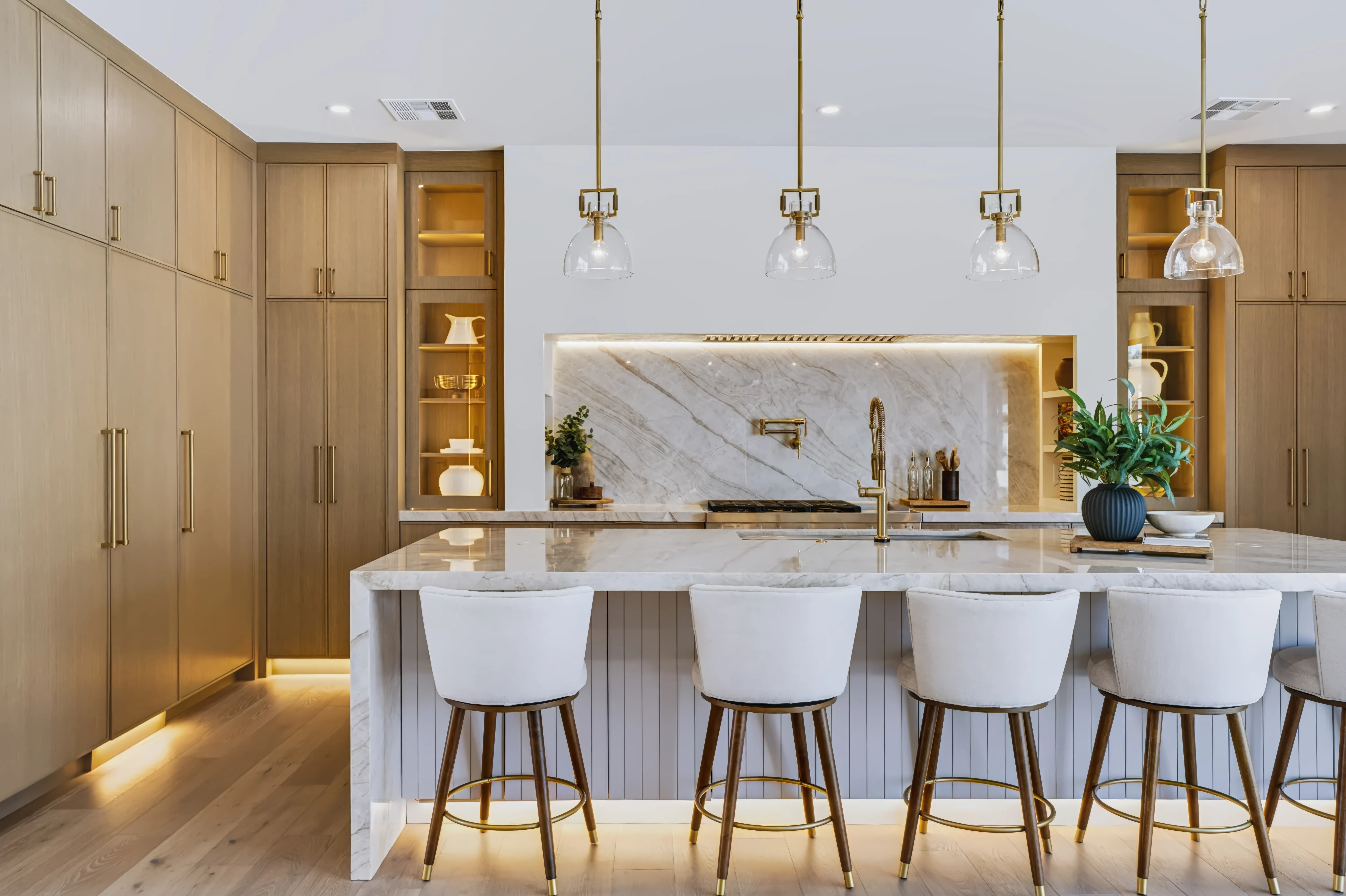 A kitchen with a waterfall marble island, brass pendants and a book-matched stone slab backsplash