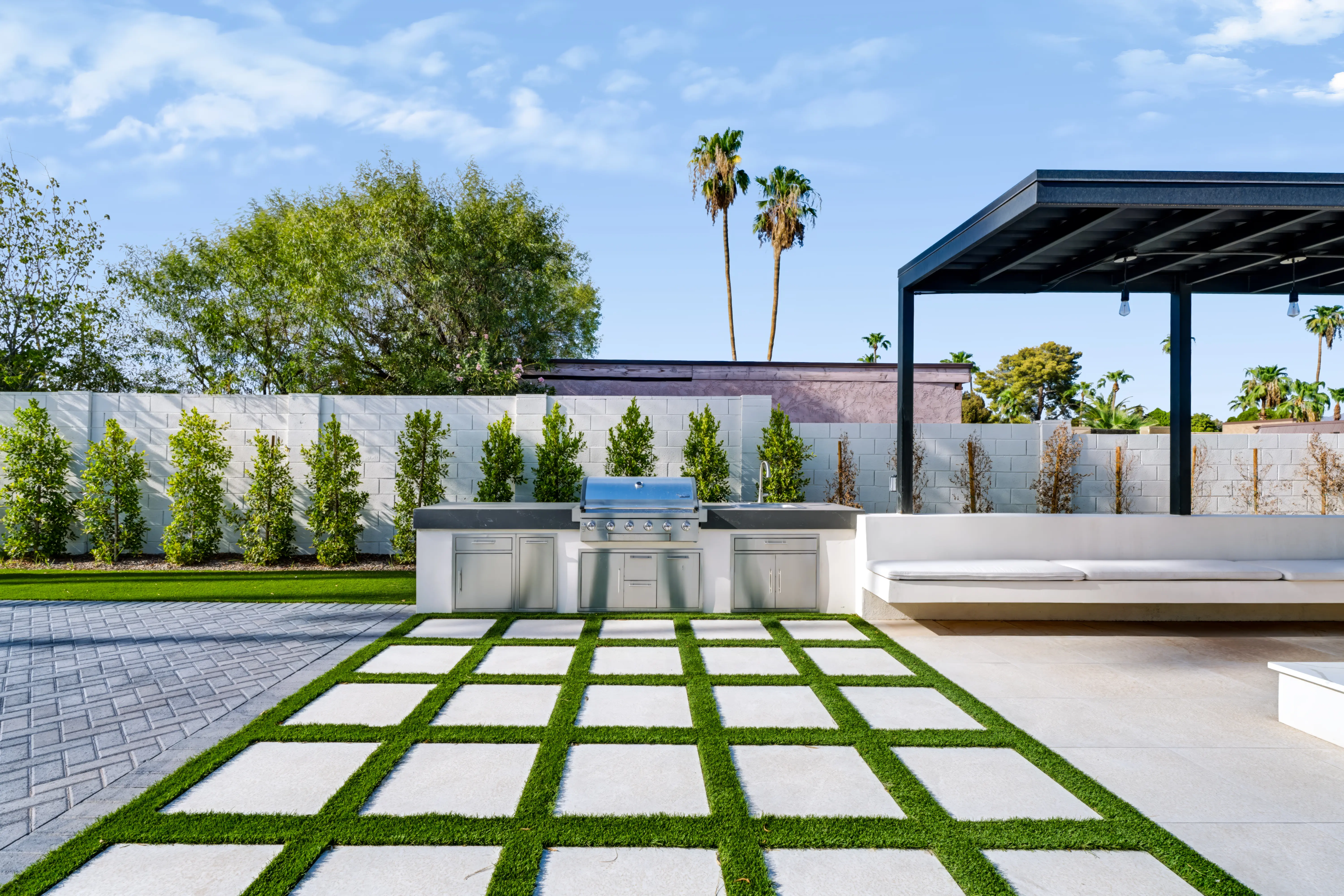 Stainless outdoor kitchen against a landscaped privacy wall with a black steel ramada and built-in bench seating