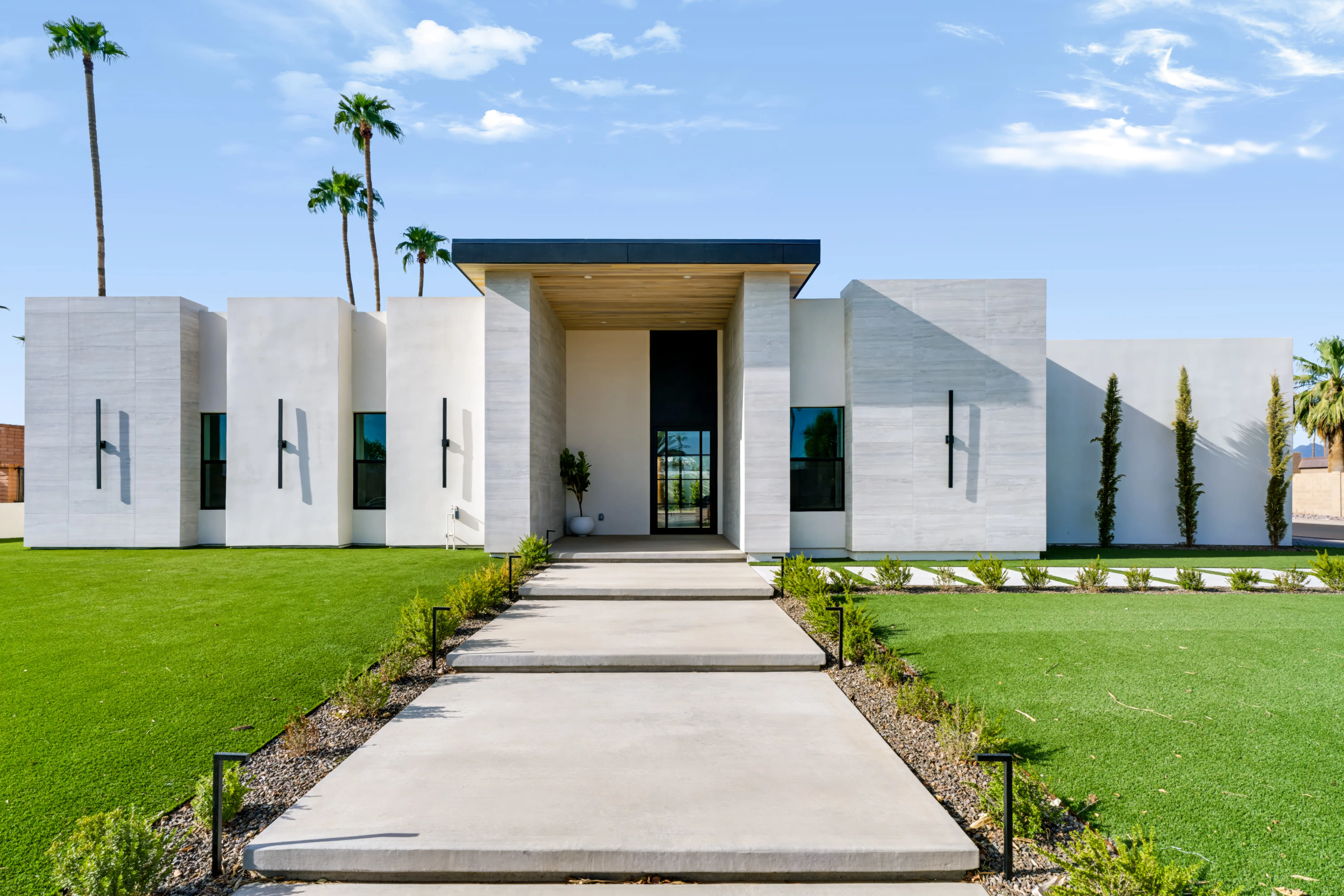Low contemporary home in afternoon sun, offset travertine and stucco volumes with a recessed black-framed entry and a clipped lawn