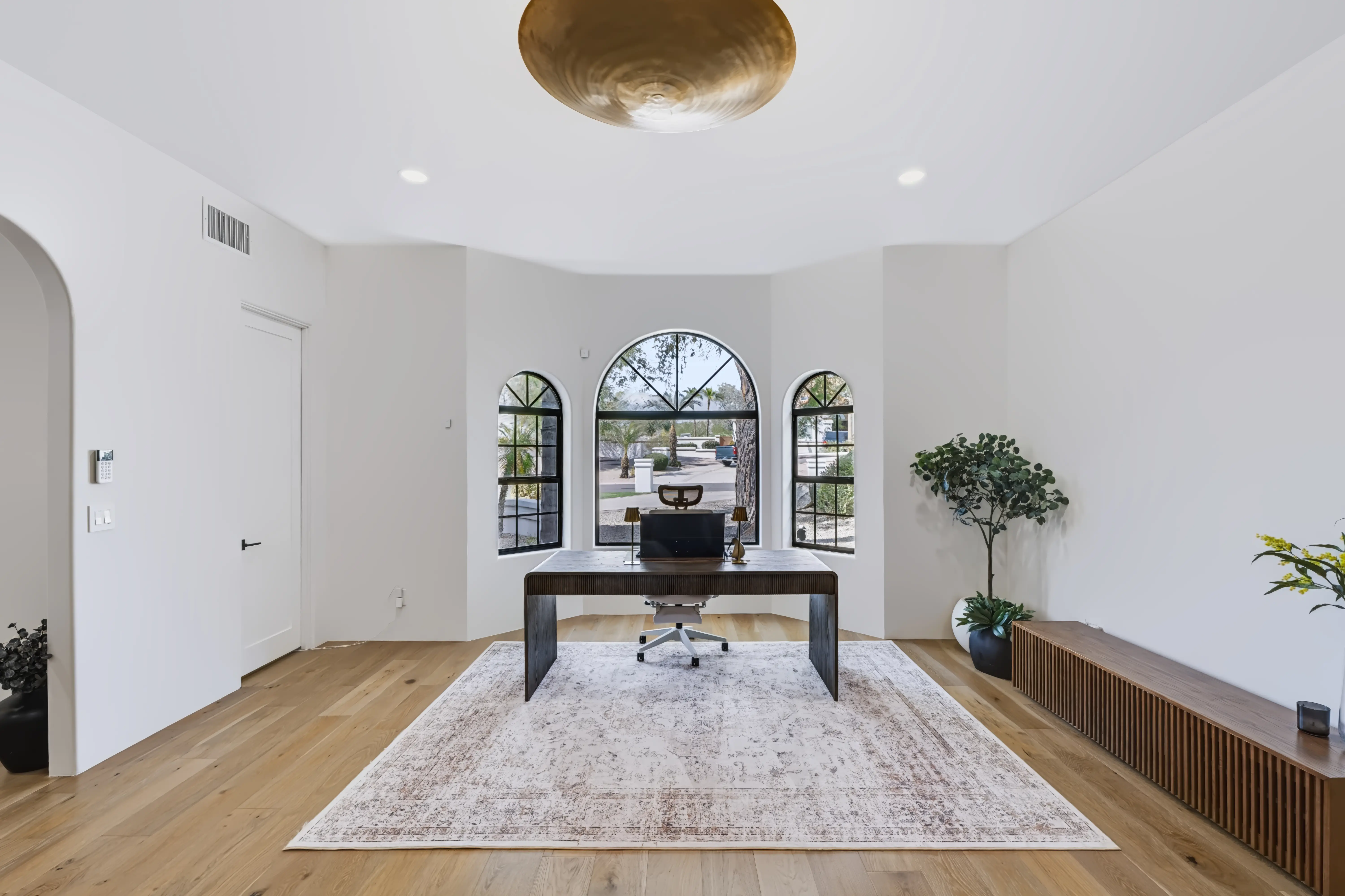 Home office in a rounded window bay, three arched steel windows behind a dark desk under a hammered-brass disc light