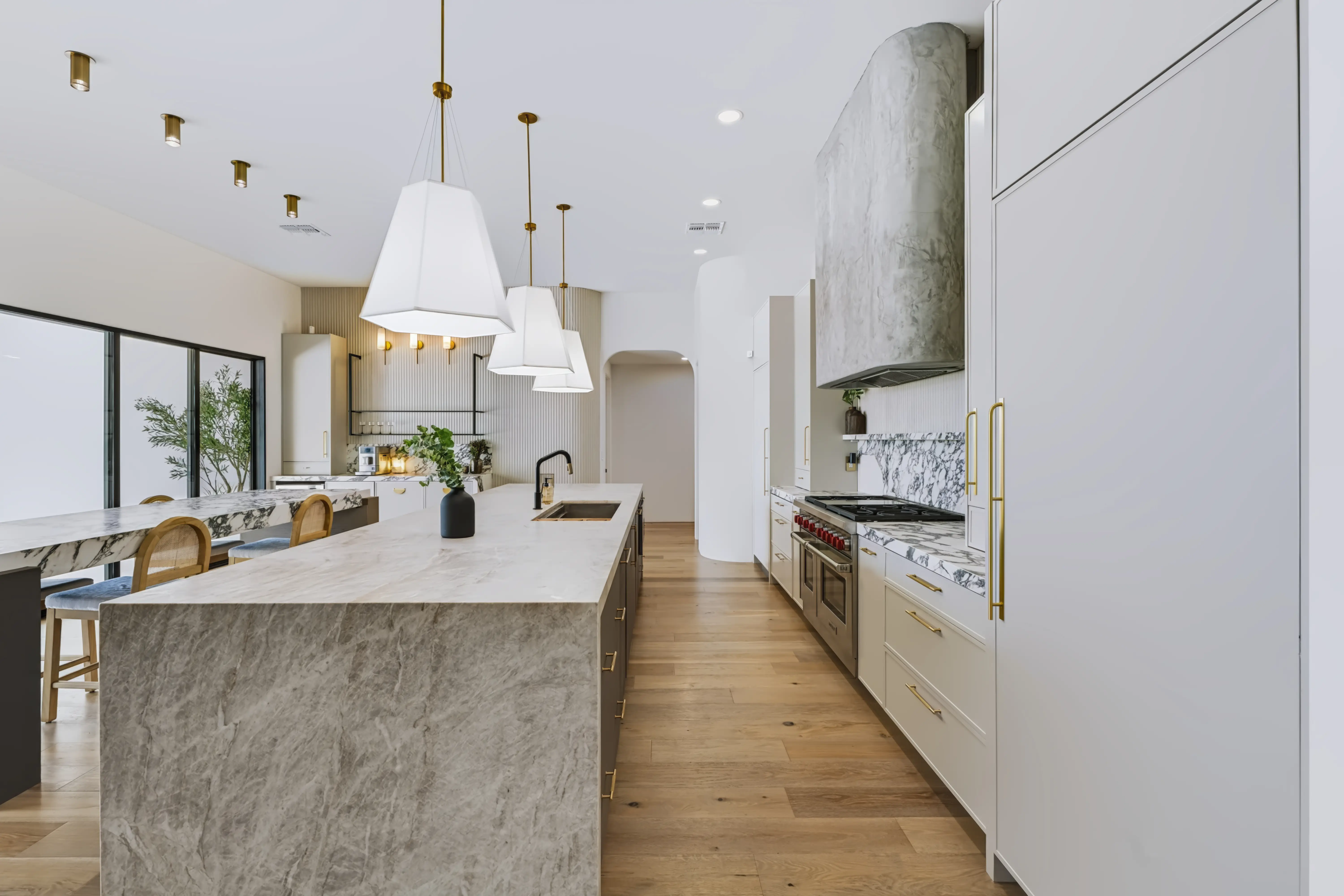 Kitchen with a long quartzite island, a plaster range hood and a marble slab backsplash under brass pendants