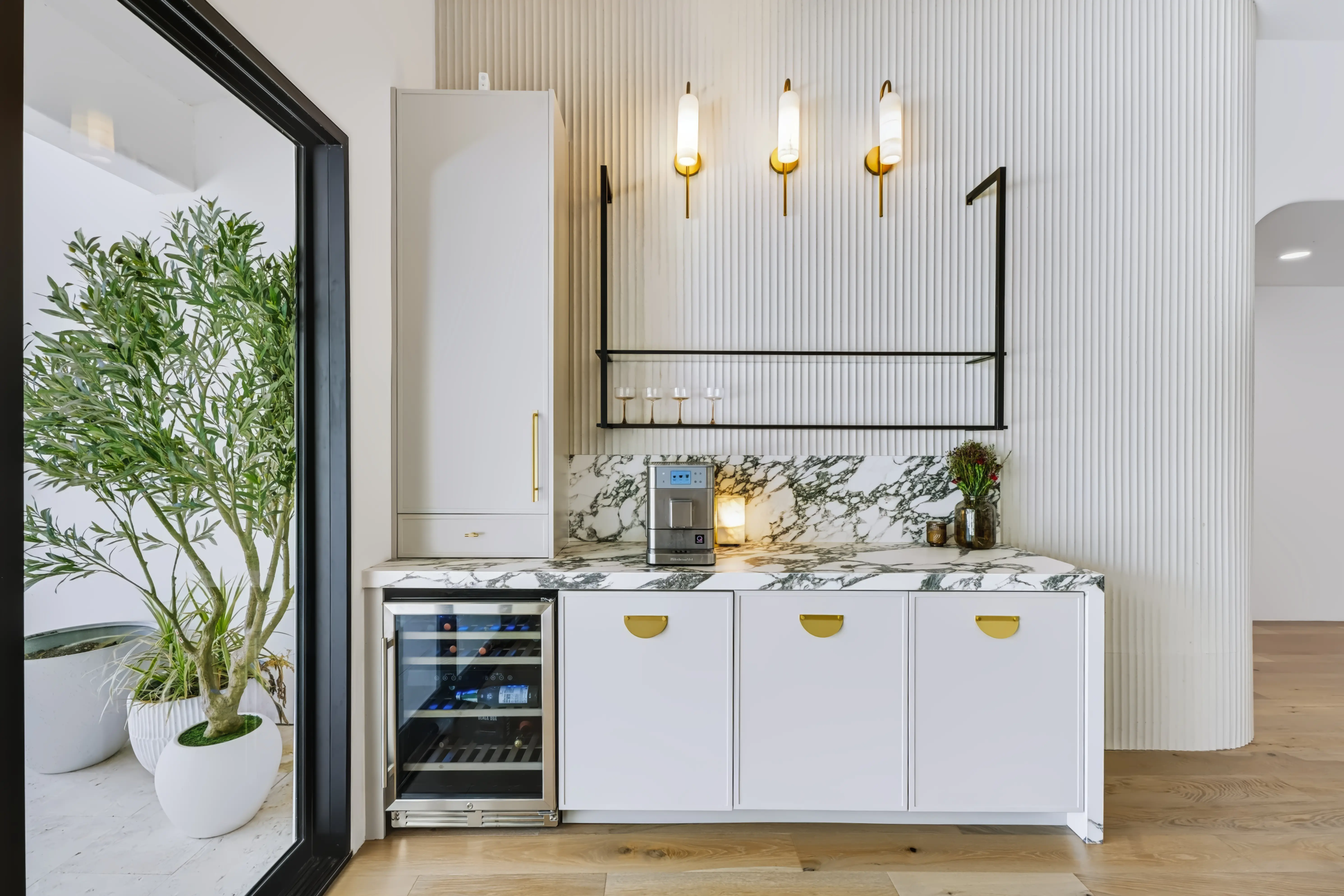 Beverage bar against a fluted wall, marble counter and backsplash, brass sconces and an undercounter wine fridge