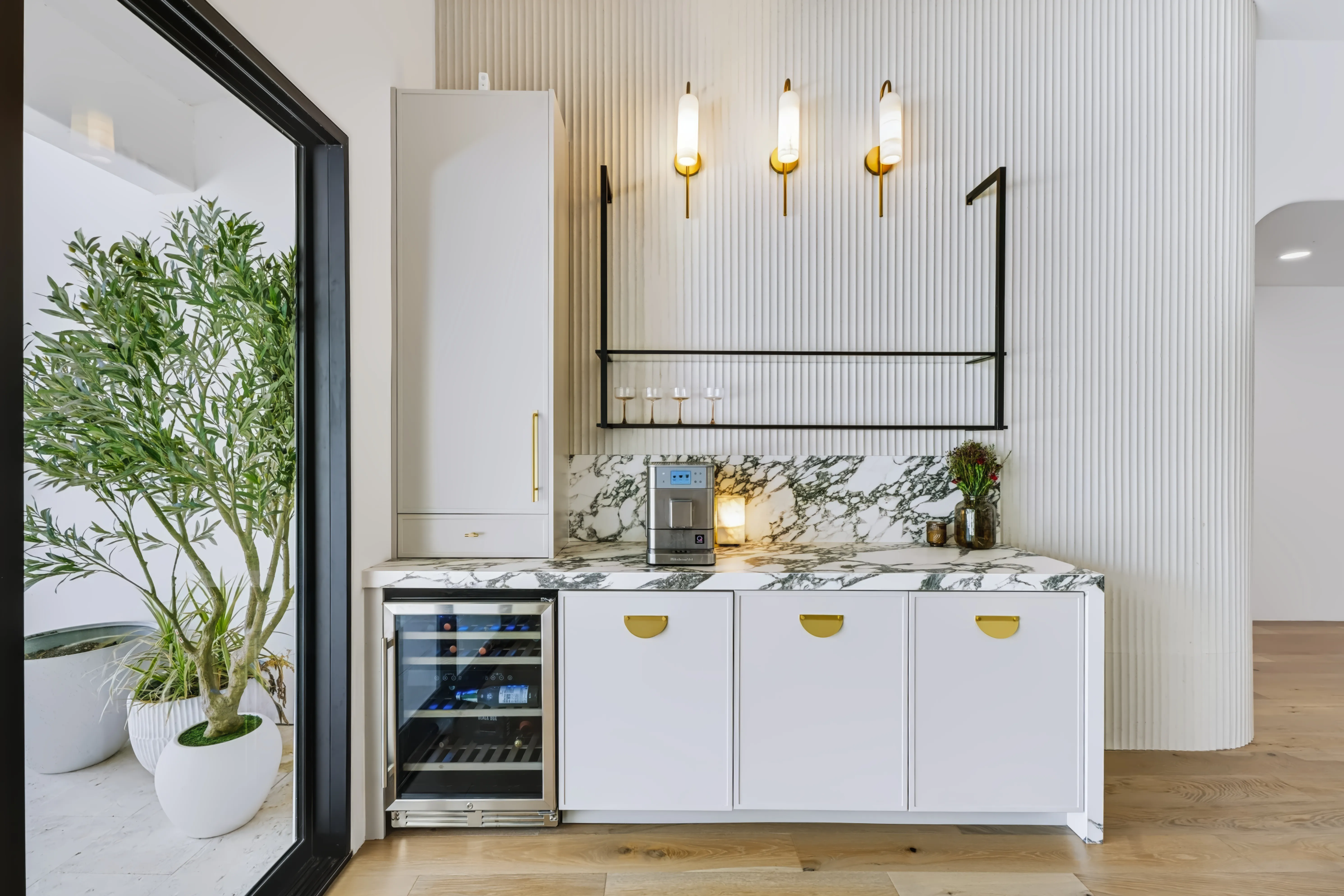 Beverage bar against a fluted wall, marble counter and backsplash, brass sconces and an undercounter wine fridge