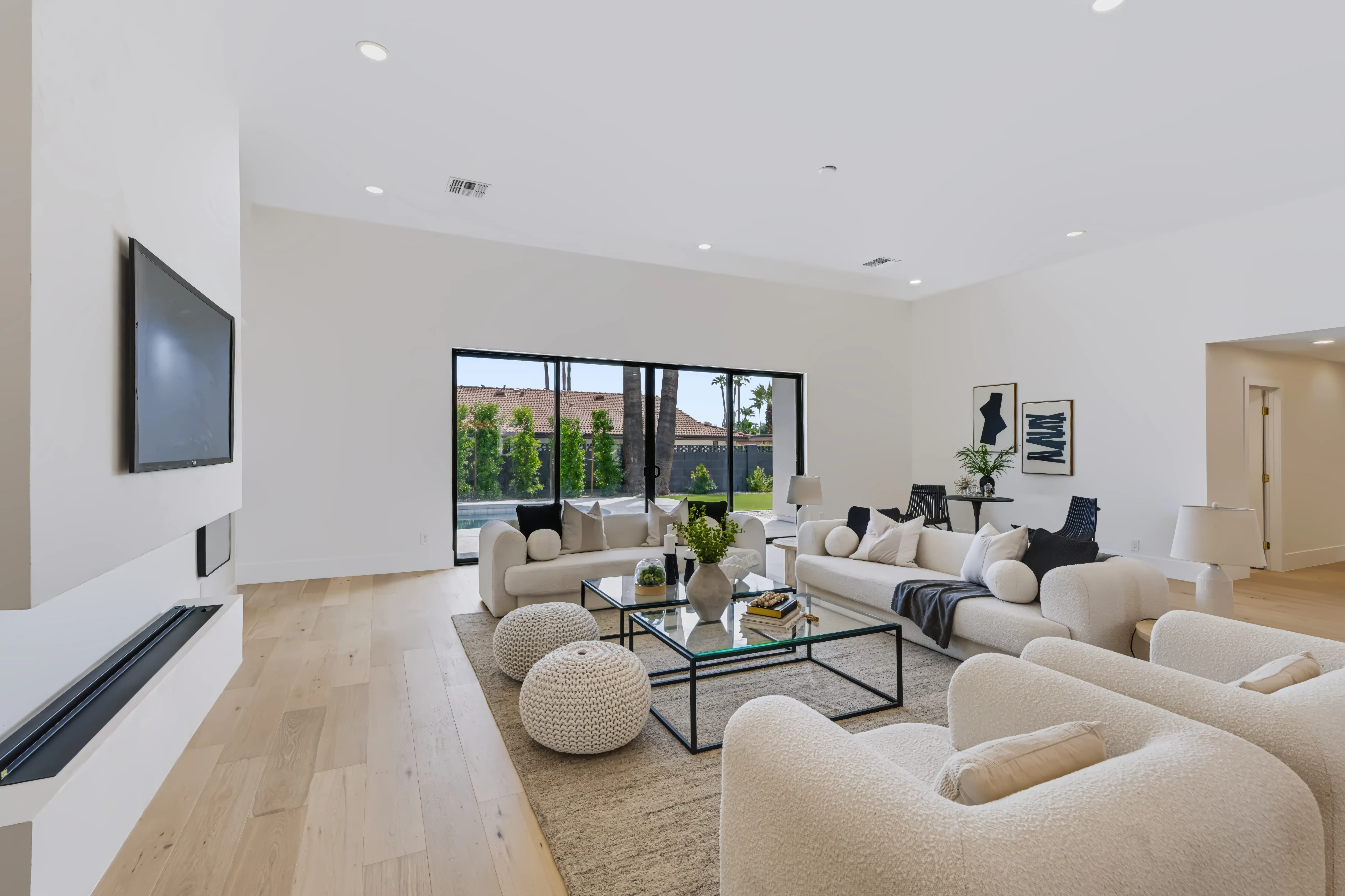 Great room in white boucle seating around a glass coffee table, a linear fireplace below the television and sliders onto the lawn