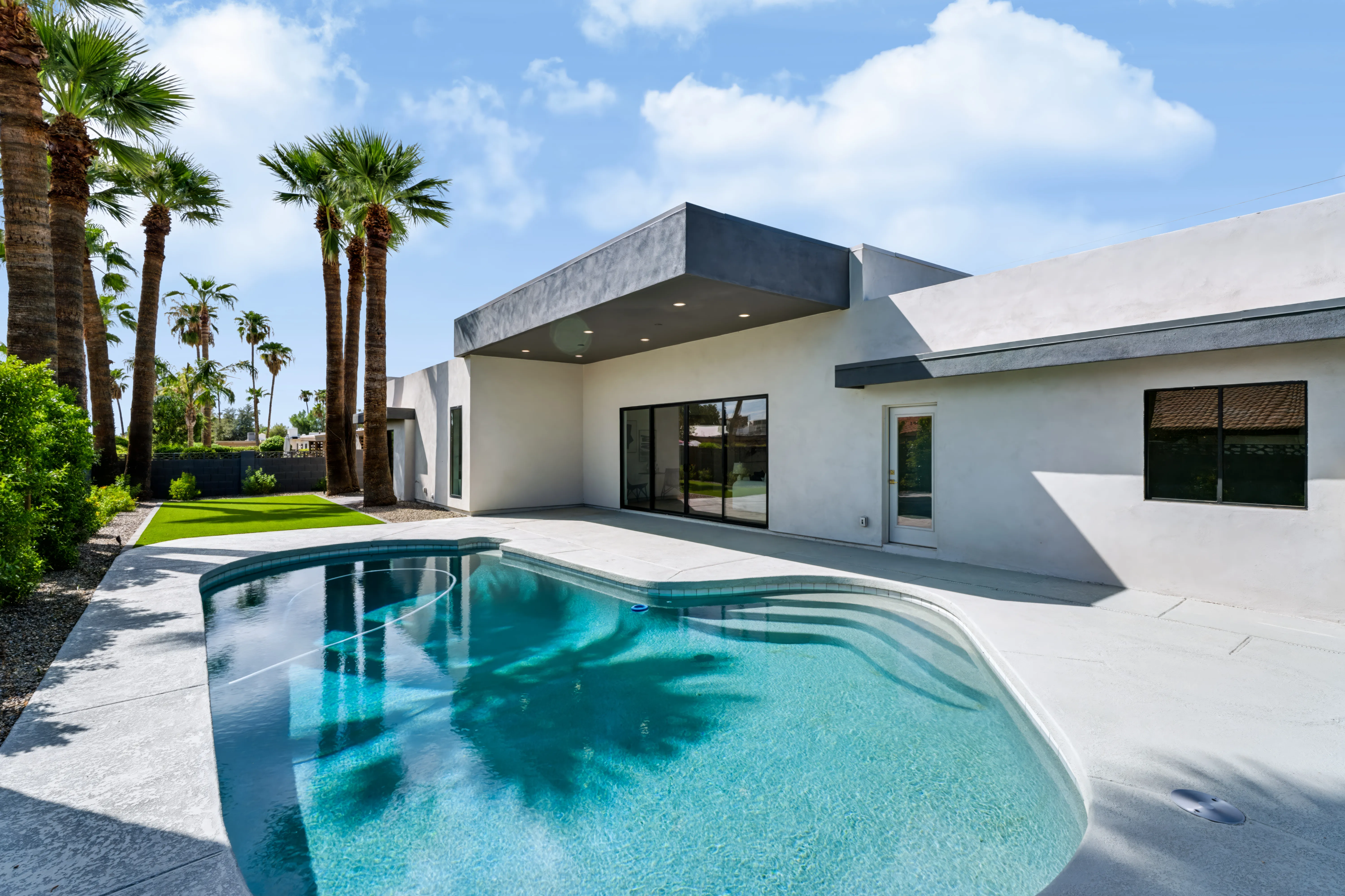 Pool from the far corner, the covered patio and sliding glass wall of the house behind a stand of palms