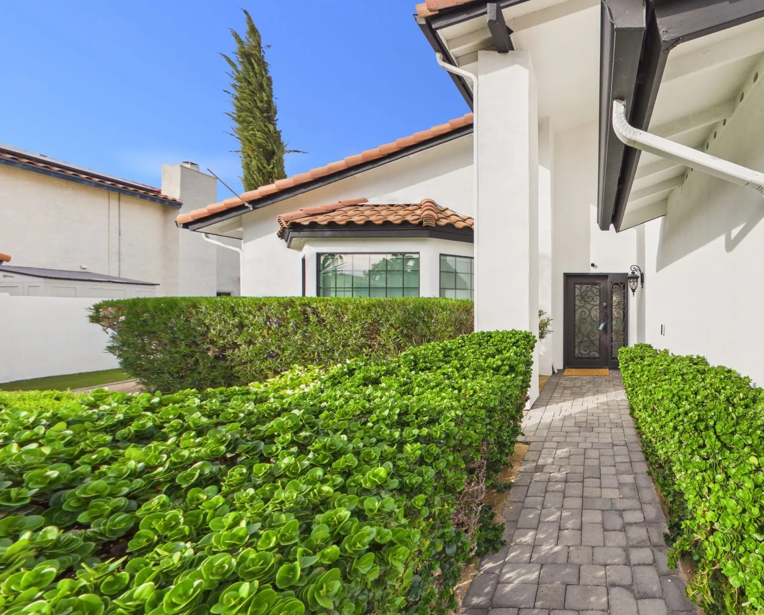Paver walk between clipped hedges to a wrought-iron front door, white stucco and a red tile roof over a bay window, a cypress behind